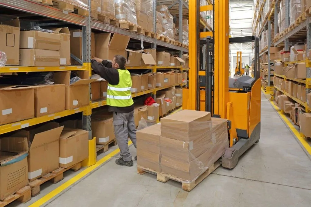 A worker handles boxes on pallets next to a reach truck in a warehouse aisle