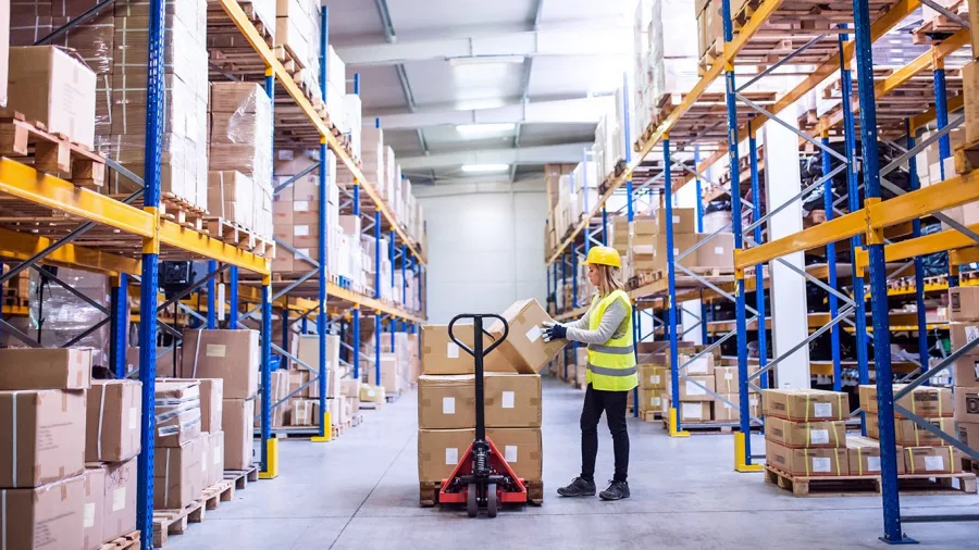 A worker pulls a pallet truck along a long warehouse aisle